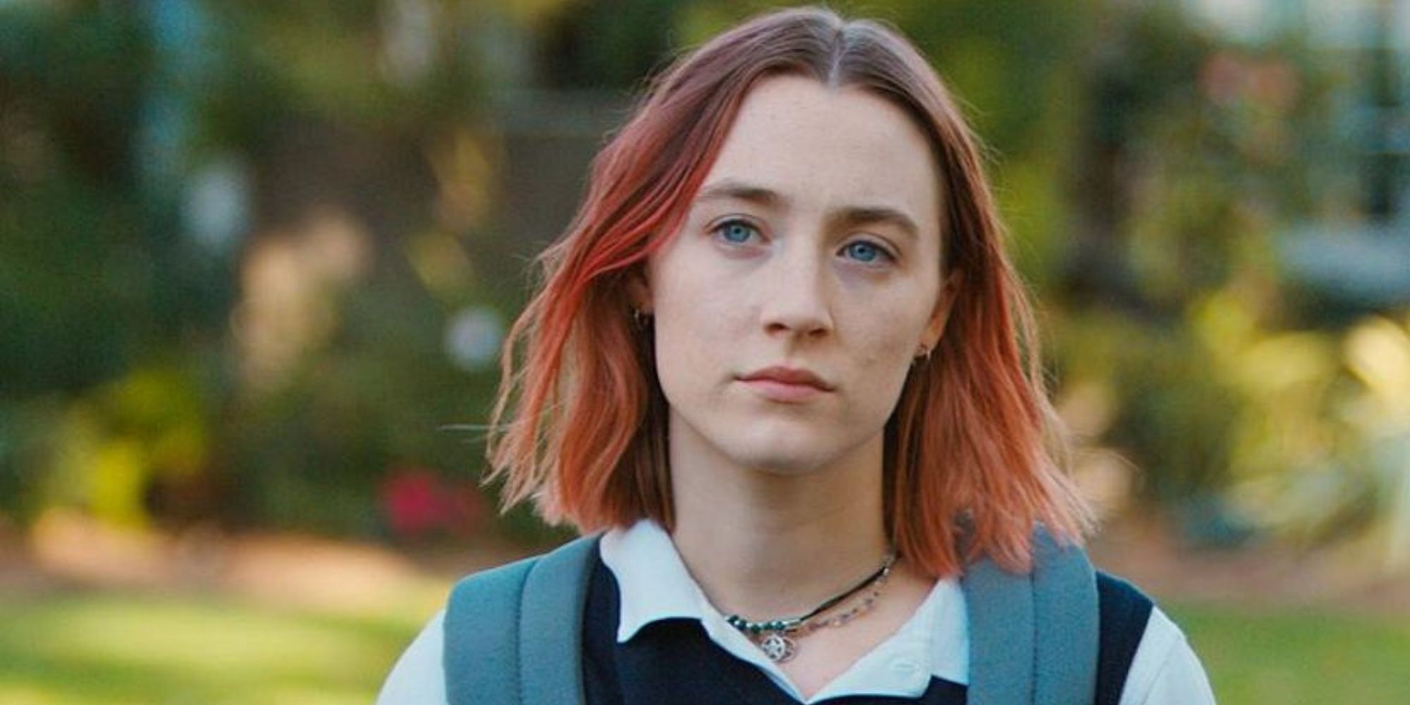 Lady Bird stands in an open space with trees in the background in Lady Bird.