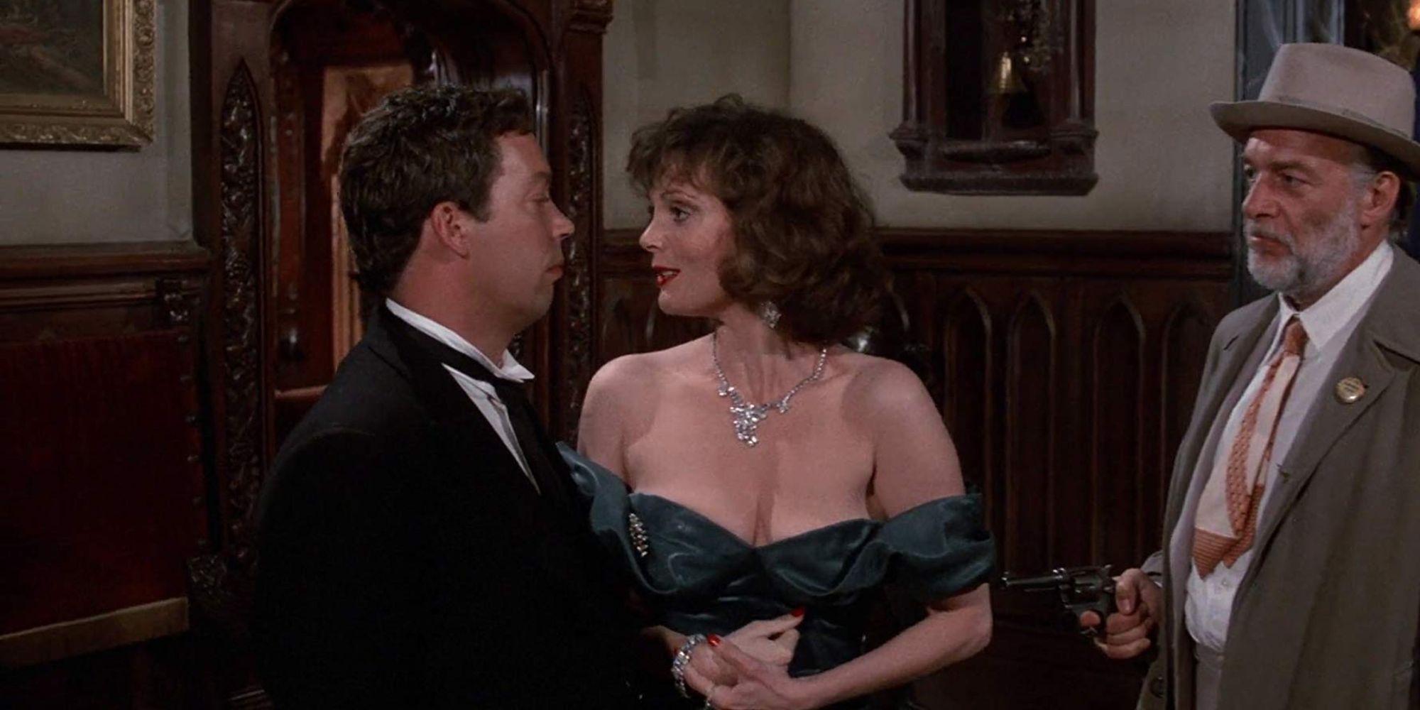 A man in a brown suit aims a gun at a man in a black suit, a woman in a blue dress stands between them.